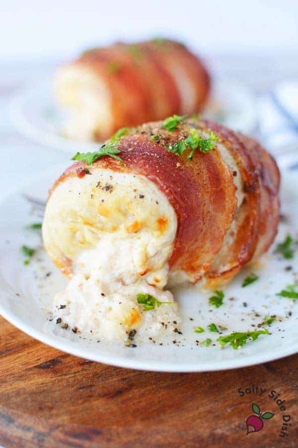 Front view of a cooked bacon-wrapped in chicken breasts with cream cheese pouring out of it. A garnish of fresh herbs