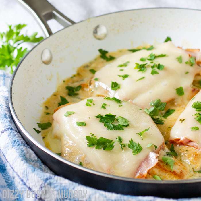 side view of cooked chicken topped with ham and melted cheese on a pan with creamy sauce