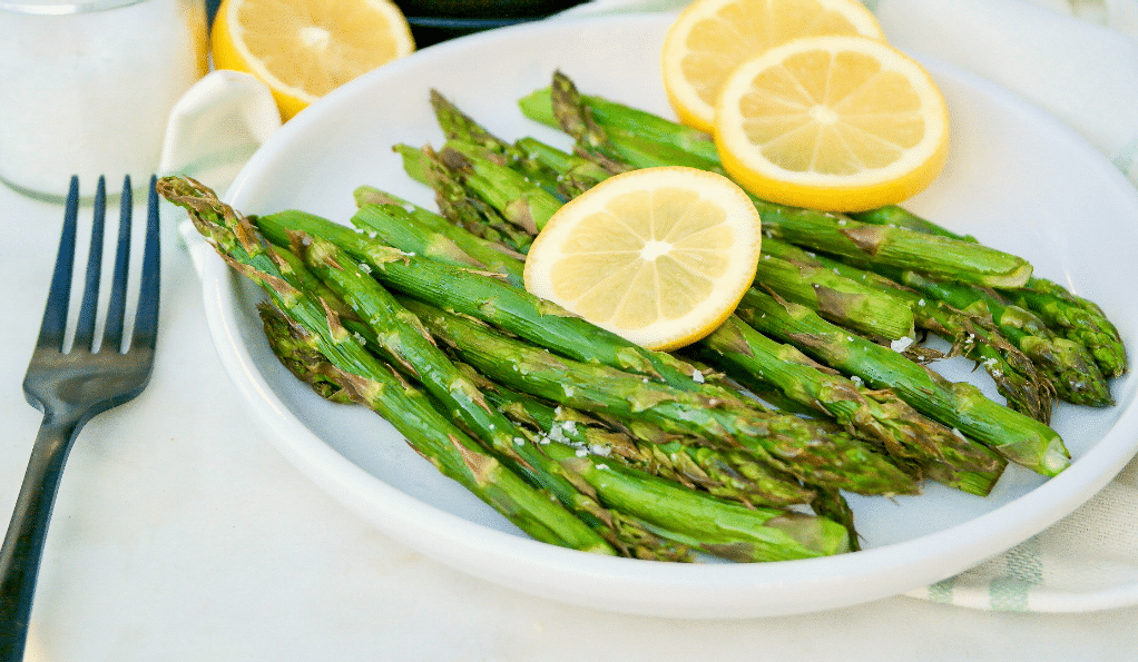 A white plate with cooked asparagus topped with bright yellow lemon slices. 