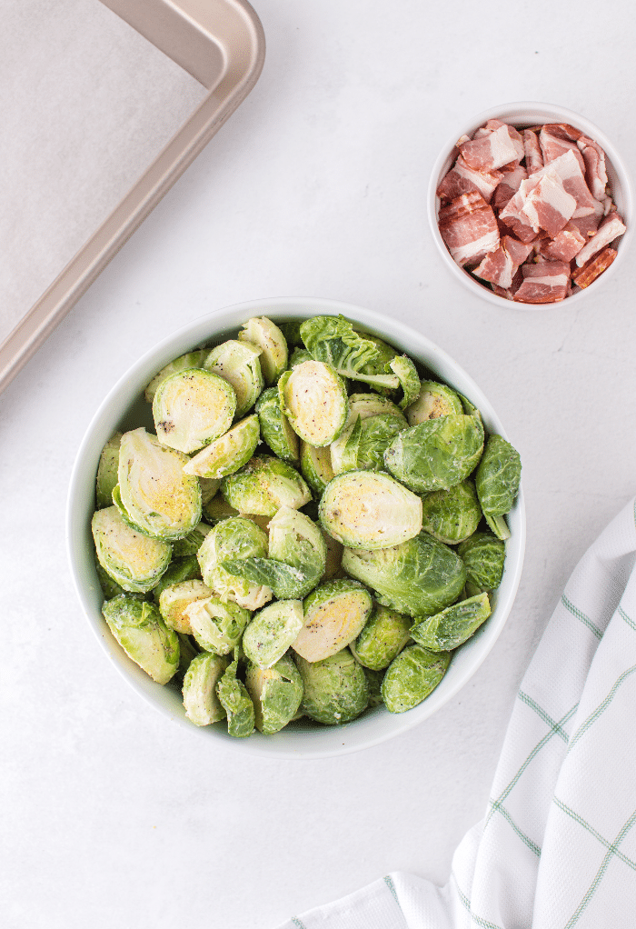 Diced bacon in a small bowl with clean and seasoned fresh Brussels sprouts ready to cook in the oven.