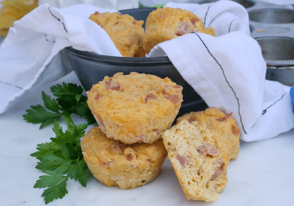 Two savory muffins on top of each other with one partially eaten on the side in from of a bowl full of muffins. 