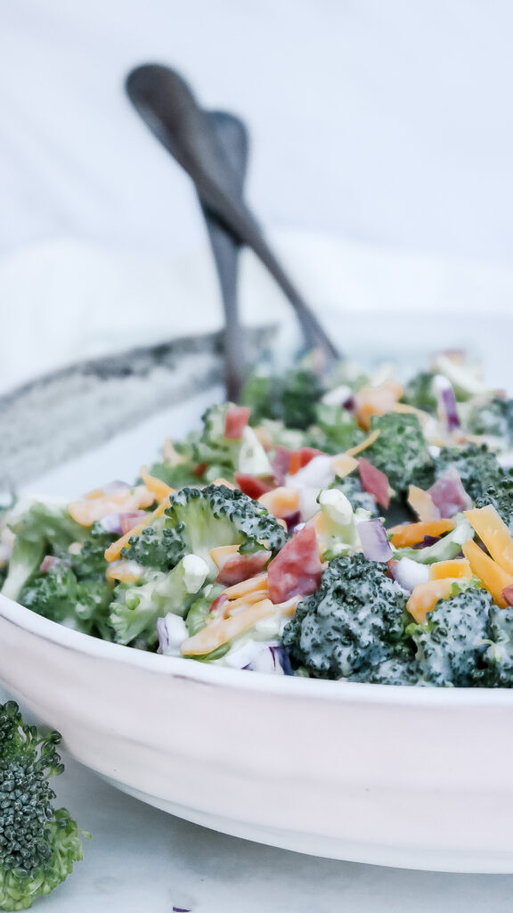 Keto broccoli salad with bacon in a white ceramic bowl with serving utensils ready to serve. 