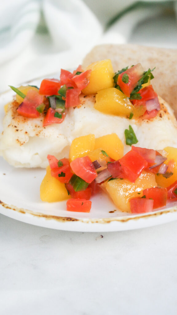 White cooked fish on a white plate topped with mango pico de gallo. 