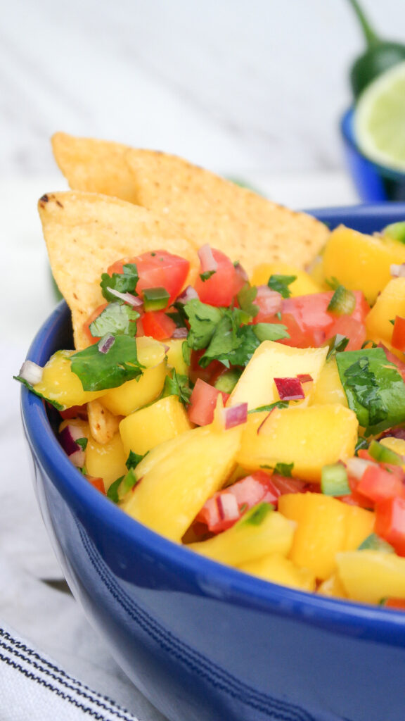 Homemade mango pico de gallo in a blue serving bowl with tortilla chips on the side. 