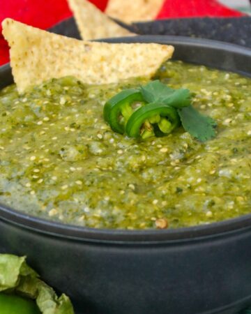 Green salsa in a black bowl with a tortilla chip and slice of jalapeno.