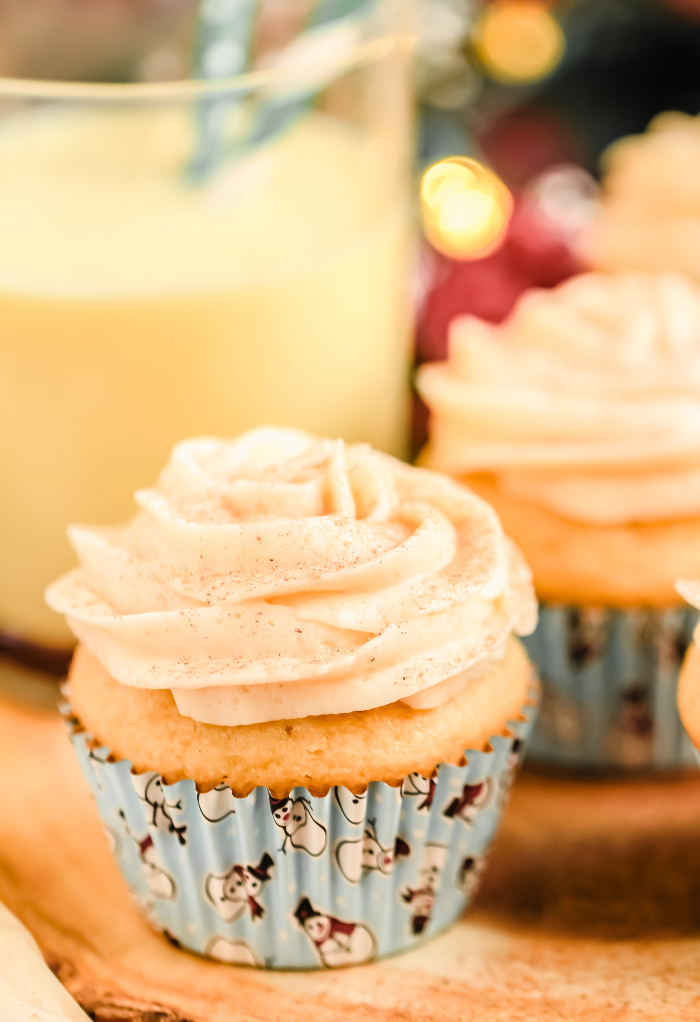 Eggnog cupcakes on a platter with a cup of eggnog behind the cupcake.