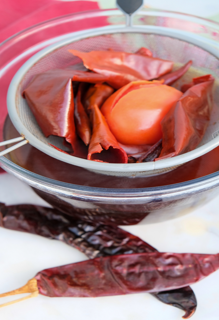 Drained cooked chile pods and tomato over a clear glass bowl. 