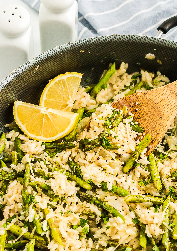 Risotto skillet dish with fresh asparagus and lemon with a wooden spoon grabbing a scoop. 