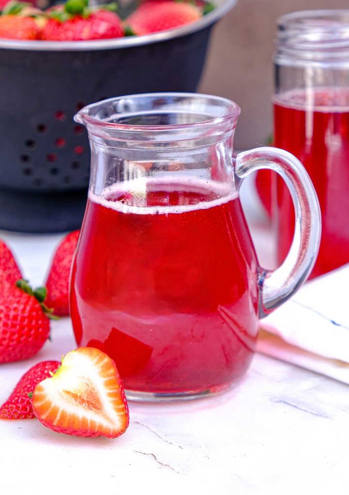 Homemade strawberry simple syrup in a clear small pitcher with fresh berries on the side.