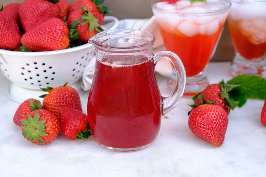 Small pitcher with strawberry syrup with a beverage in the back. 