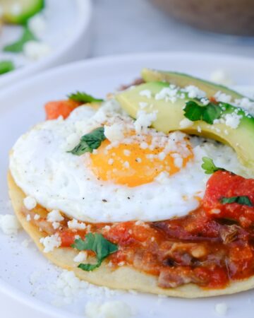 Homemade huevos rancheros with sunny side up egg and sliced avocados on a white plate.
