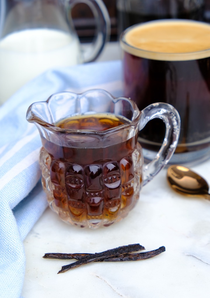 Vanilla syrup in a small pitcher next to a vanilla bean. 