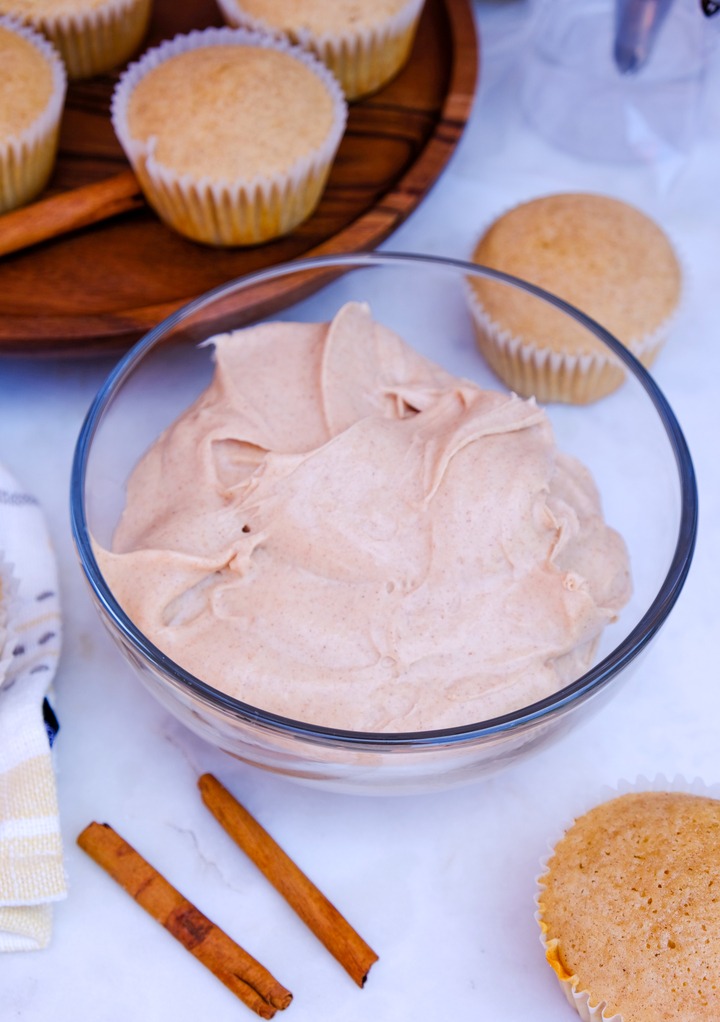 Cinnamon cream cheese frosting in a glass mixing bowl with plain cupcakes on the side. 