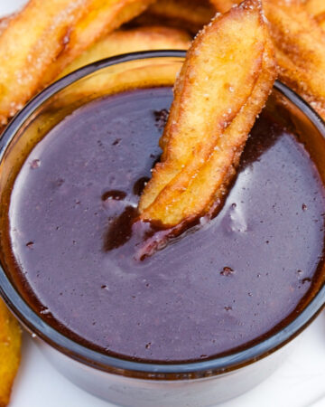 Homemade Ghirardelli chocolate sauce in a small clear serving bowl with a churro dipped in the center.