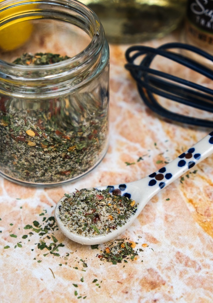 Dry spices in a clear jar with a spoon on the side with a scoop of the spices. 