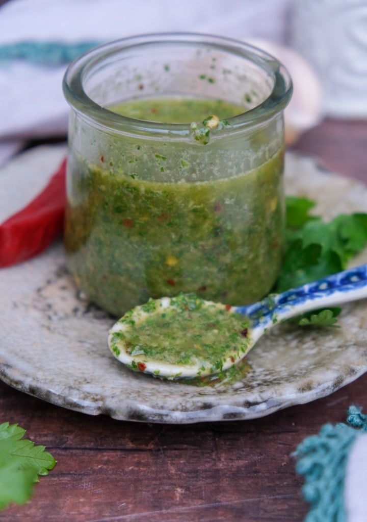 Homemade spicy chimichurri sauce in a small clear jar placed on a tan place with a spoon on the side. 