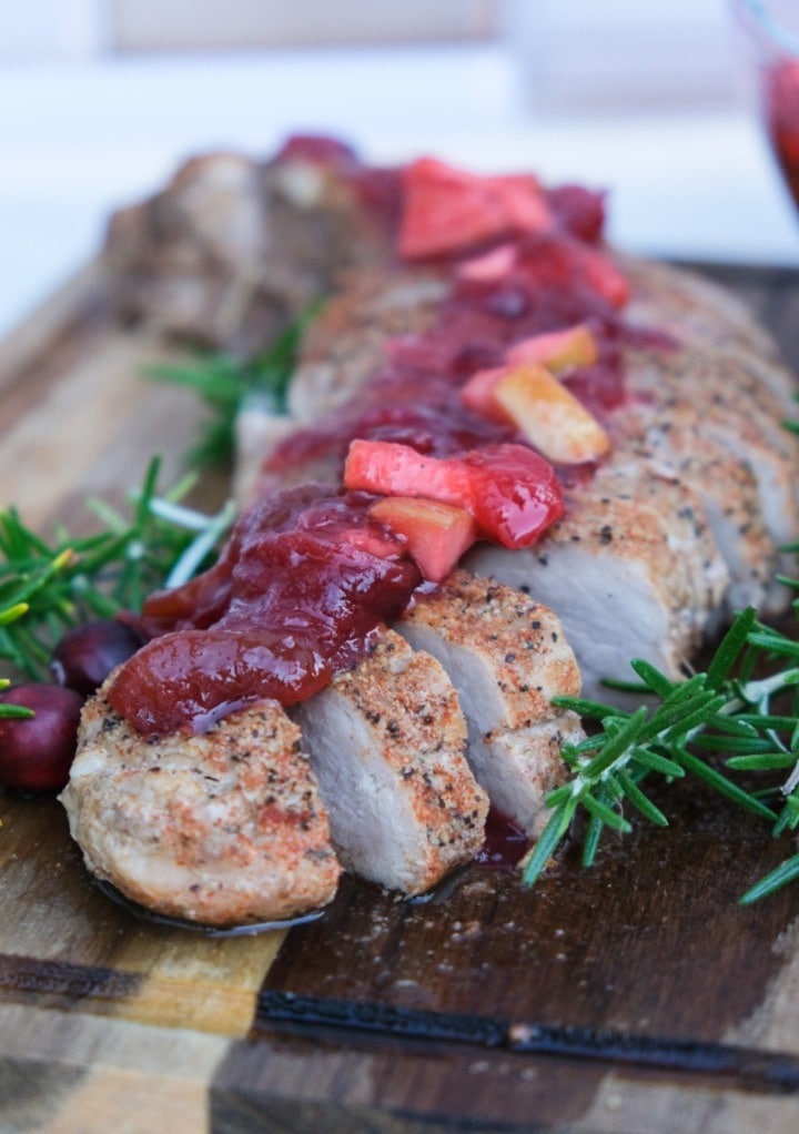 Homemade apple cranberry chutney over sliced pork on a brown cutting board. 