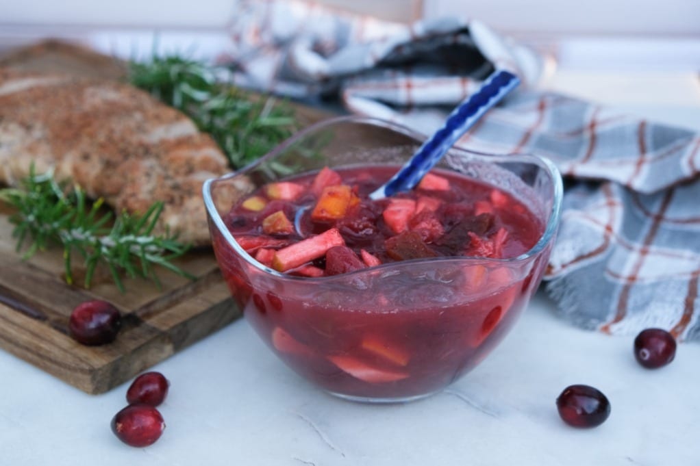 Fresh cranberry chutney in a clear serving bowl with pork in the background on a cutting board. 