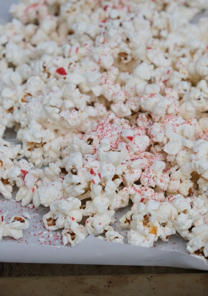 Fresh popcorn topped with white chocolate, crushed peppermint candy, and red sanding sugar on a baking pan. 