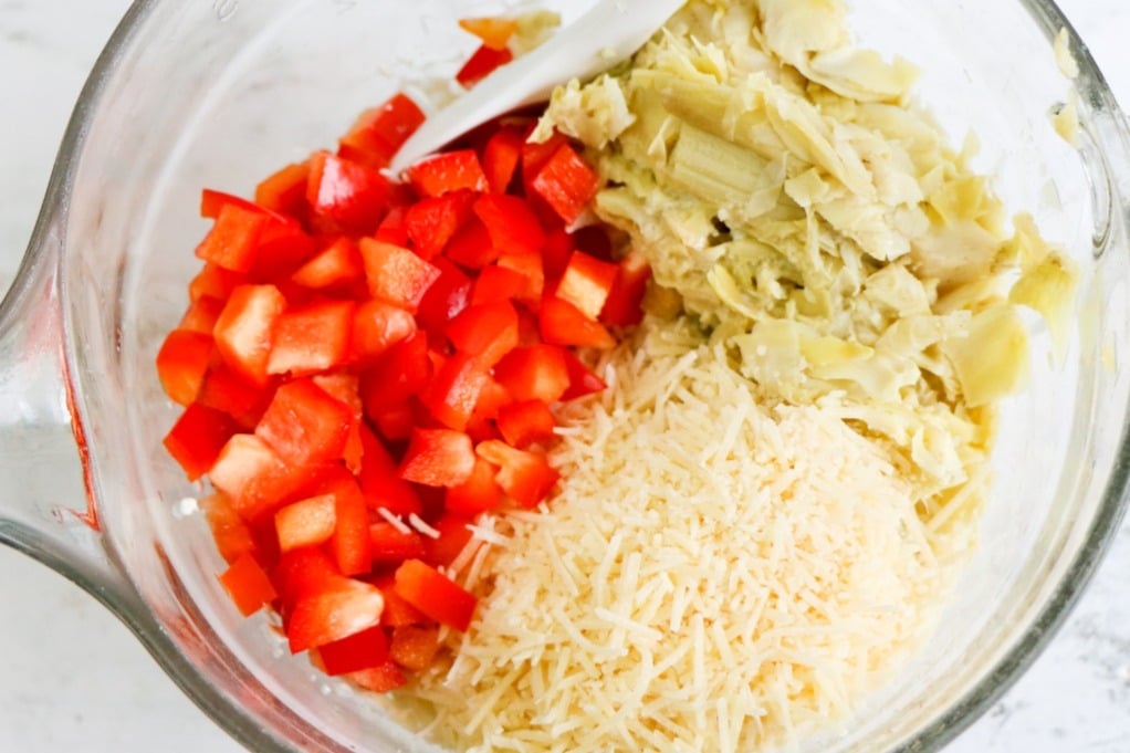 Diced red pepper, artichoke hearts and parmesan cheese in a clear mixing bowl. 