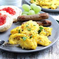 Cooked scrambled eggs on a light blue plate with sausage and a sliced English muffin.