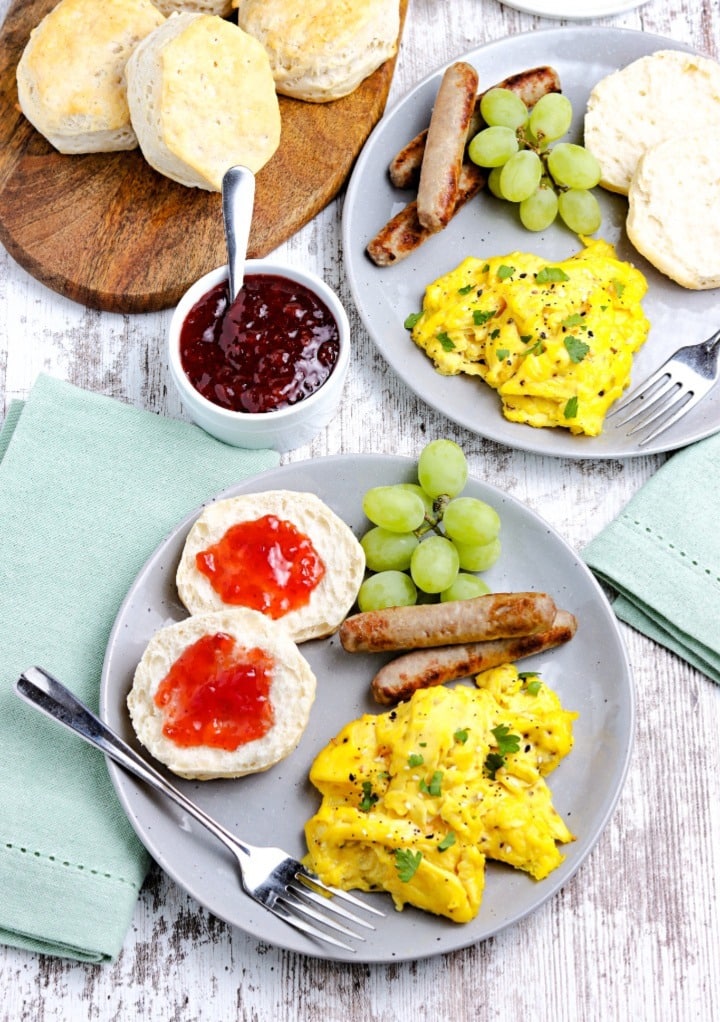 Top view of two breakfast plates with eggs, sausage and biscuits with jam. 