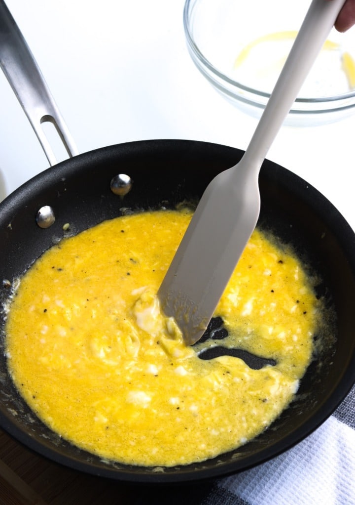 A non stick pan cooking eggs with a gray spatula pushing the eggs as they cook. 