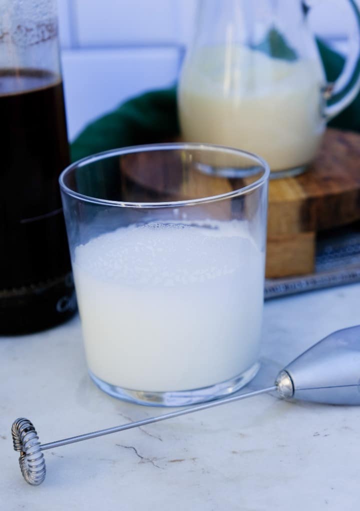 Warm milk in a clear glass with a hand frother on the side. 