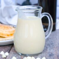 Homemade white chocolate syrup in a small clear pitcher with pancakes in the background.