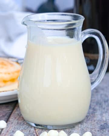 Homemade white chocolate syrup in a small clear pitcher with pancakes in the background.