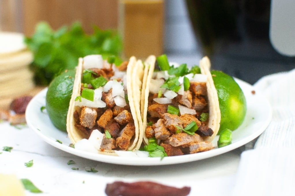 Spicy pork tacos propped with sliced limes of a plate with an air fryer on the side. 