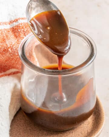 Vegan caramel sauce in a clear jar with a spoon dripping with the sauce.