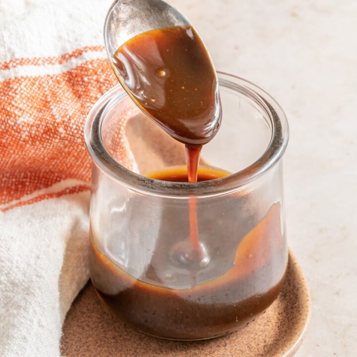 Vegan caramel sauce in a clear jar with a spoon dripping with the sauce.