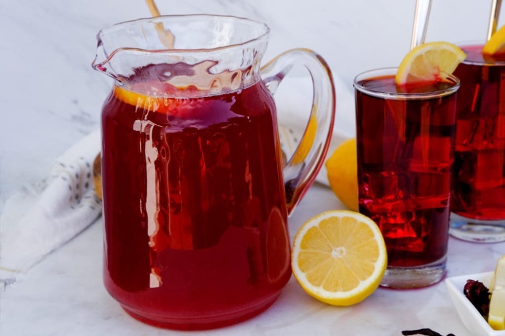 A pitcher filled with hibiscus lemonade with fresh lemon on the side. 