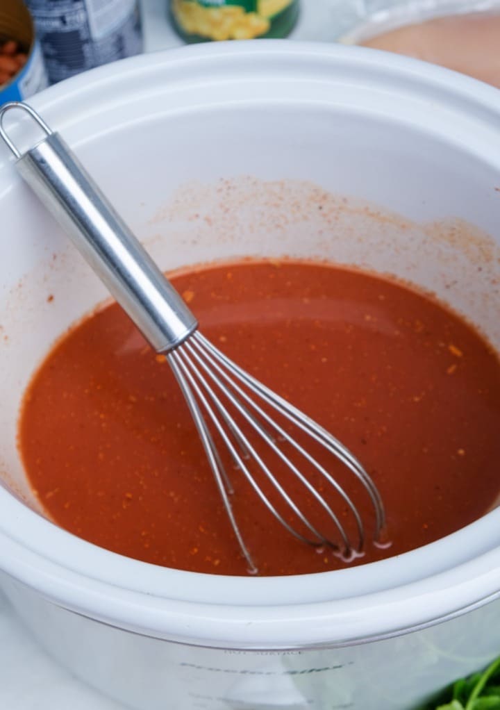 Tomato sauce and spices added to a white slow cooker with a whisk mixing the ingredients. 