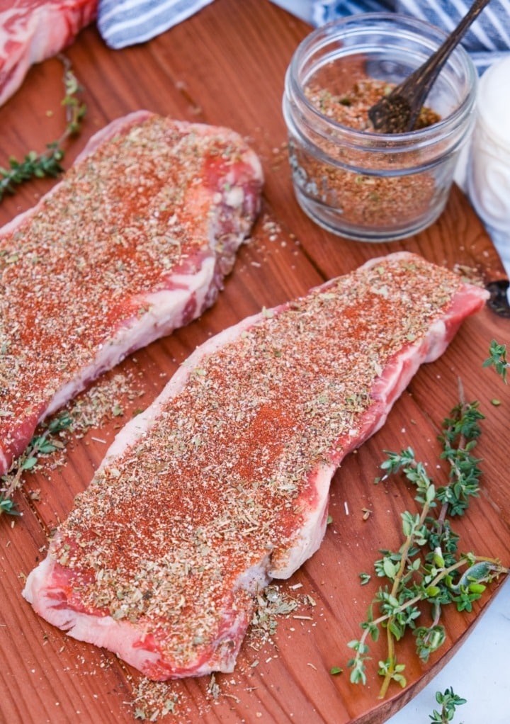 Raw steaks on a wooden cutting board with homemade steak seasoning mix before cooking.