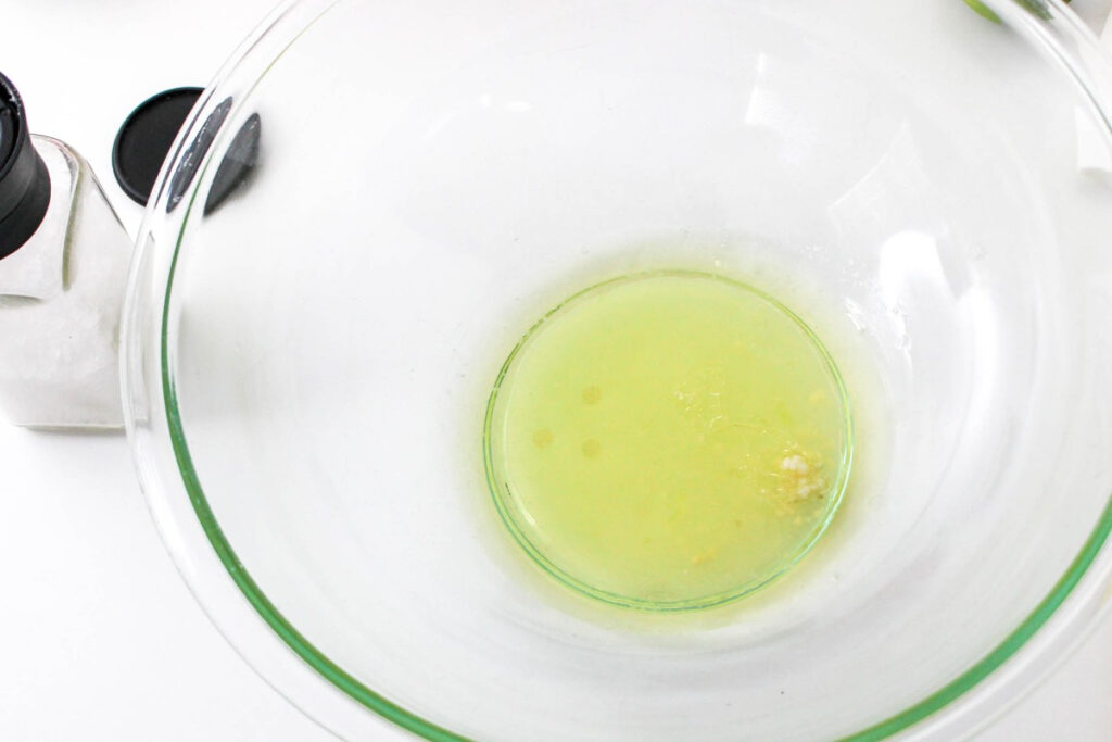 Homemade dressing in a clear mixing bowl with olive oil, garlic and lime juice. 