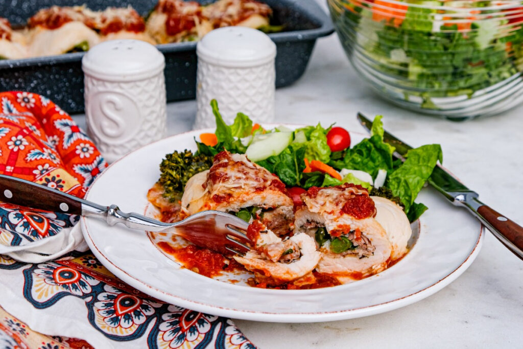 A white dinner plate with chicken rolled with broccolini and prosciutto topped with marinara sauce. 