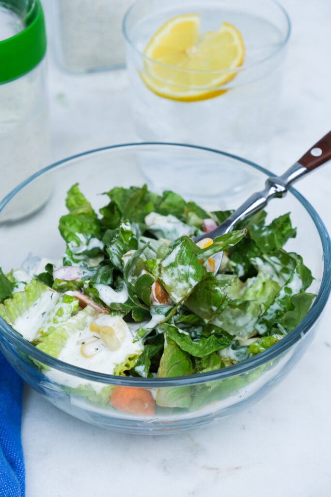 Clear bowl with salad and a fork ready to eat with a glass of lemon water in the background. 