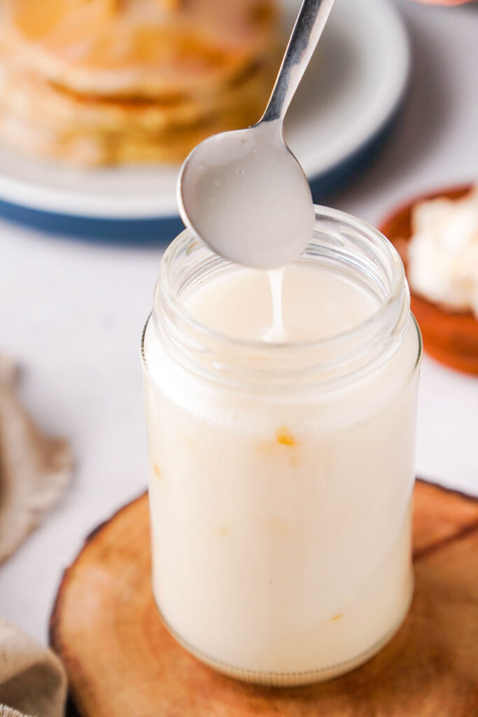 Homemade coconut syrup in a clear jar with a spoon drizzling the sryup.