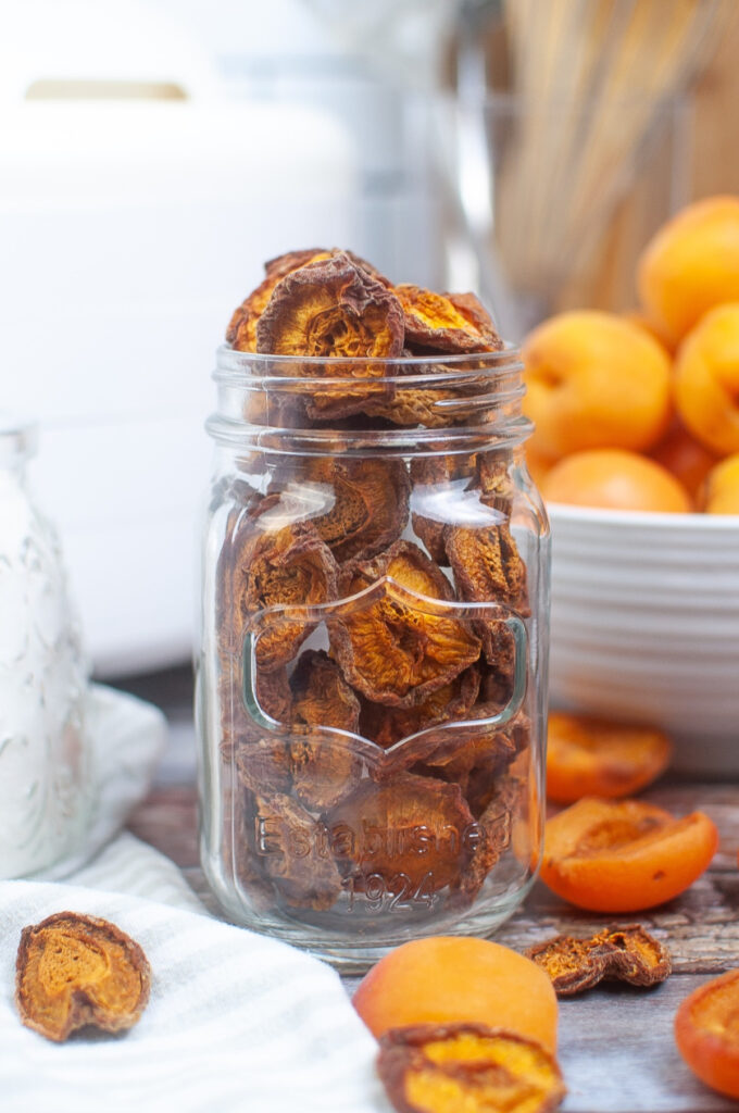 Dried apricots in a clear tall mason jar with fresh apricots in a bowl on the side. 