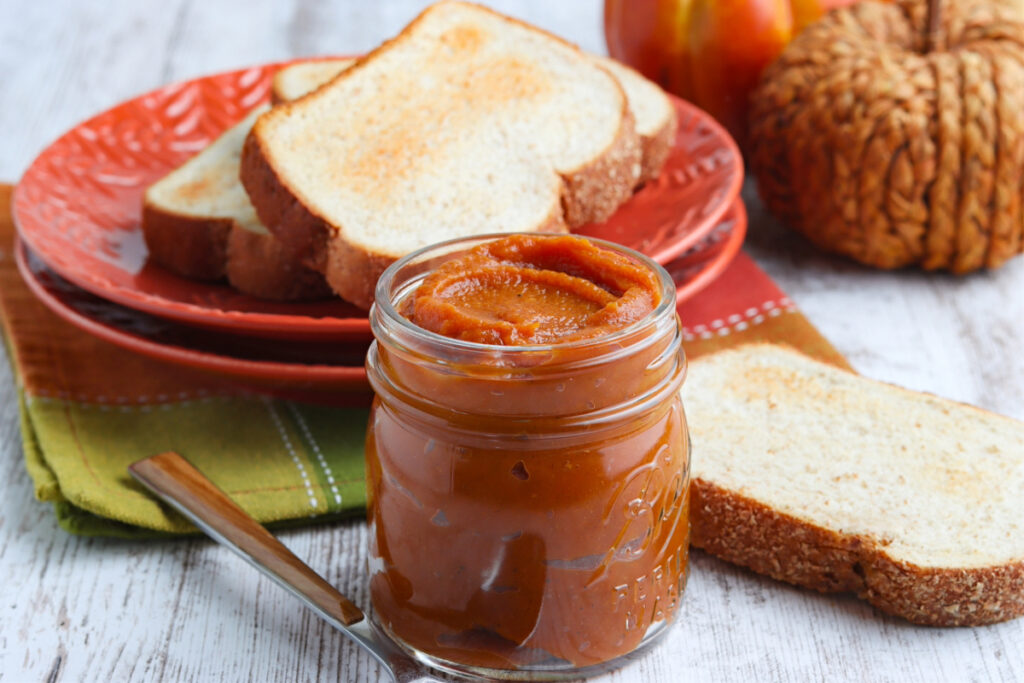 Homemade cooked pumpkin butter in a glass jar with a plate on the side topped with toast. 