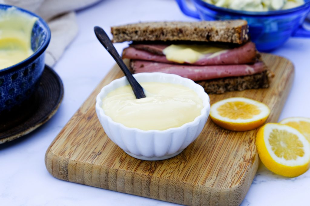 A small white condiment bowl with mayonnaise on a tan cutting board with a half sandwich on the side. 
