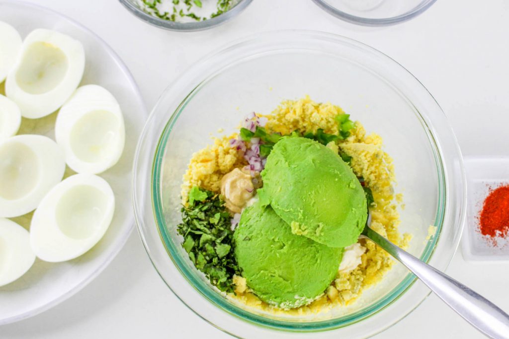 Deviled egg ingredients with avocado in a clear mixing bowl. 