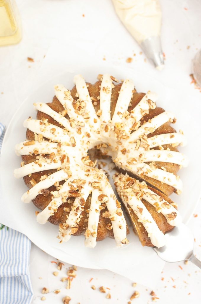 Overhead view of a carrot cake bundt cake on a white serving platter with a slice ready to be placed on a plate.