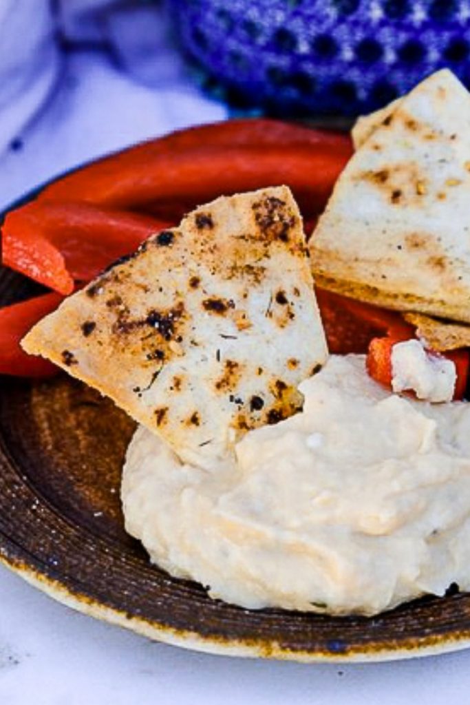 Small brown plate with a scoop of hummus and a pita chip.