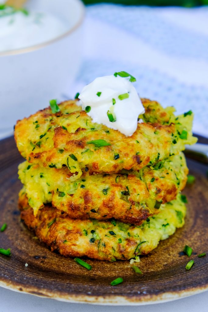 A stack of three zucchini fritters topped with sour cream on a brown plate.