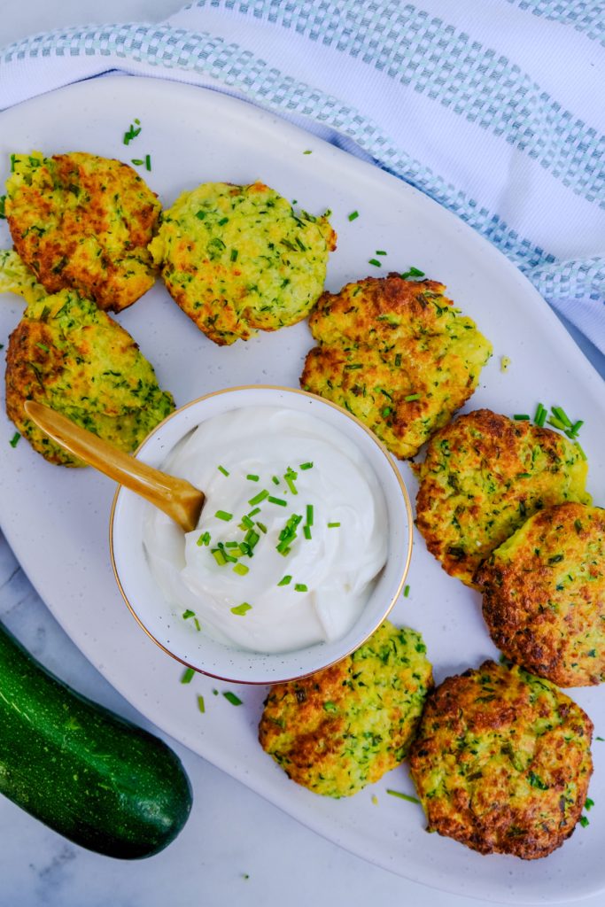 Overhead view of zucchini fritters on a white serving plate with sour cream in the center.