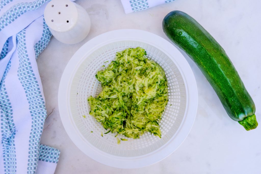 Grated zucchini in a white colander draining excess water. 