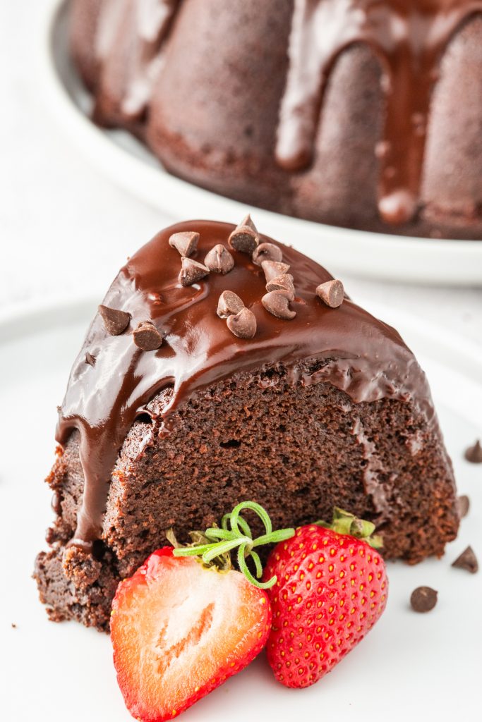 Chocolate bundt cake with chocolate glaze on a white plate with a sliced strawberry on the side. 